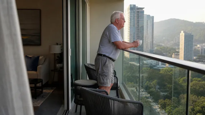 A retired man on a balcony looking out over green hills and towers